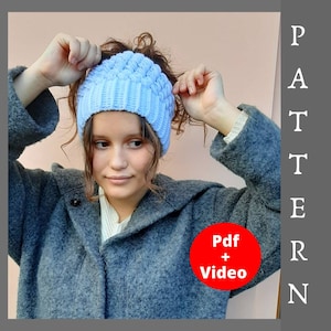 May include: A light blue crochet hat with a bun hole. The hat is being worn by a person. The text "Pdf + Video" is on a red circle.