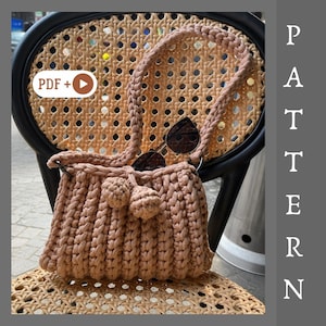 May include: A light brown, hand-knitted handbag with a braided strap and two decorative pom-poms. The bag is resting on a wicker chair. Sunglasses are tucked inside the bag. The word "PATTERN" is visible in the image.