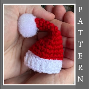 Può includere: Un piccolo cappello di Babbo Natale all'uncinetto con un pon pon bianco e strisce rosse e bianche.