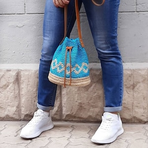 May include: A blue and beige crocheted bucket bag with a brown leather strap. The bag is worn over the shoulder of a person wearing blue jeans and white sneakers.