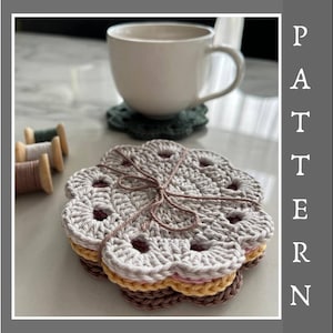 Może przedstawiać: Stos ręcznie robionych podkładek szydełkowych w odcieniach szarości, żółci i brązu, związanych sznurkiem. Biały kubek stoi na zielonej podkładce, z szpulkami nici w tle. Słowo "PATTERN" jest wyświetlane pionowo.