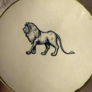 May include: A round, off-white ceramic plate featuring a detailed black ink illustration of a standing lion in the center. The plate has a thin, dark-colored rim that frames the circular edge, providing a clean and simple aesthetic for the dish.