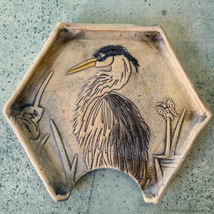 May include: Hexagonal ceramic dish with a raised relief of a heron. The bird has a yellow beak and is depicted with detailed feathers. The dish is beige with blue accents and a cutout on one side. The heron is surrounded by stylized plants.