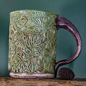 May include: A ceramic mug featuring a raised, textured surface with a repeating pattern of green monstera and fern leaves. The mug has a dark brown, curved handle that resembles a tree branch and a base with a matching brown, earthy finish.