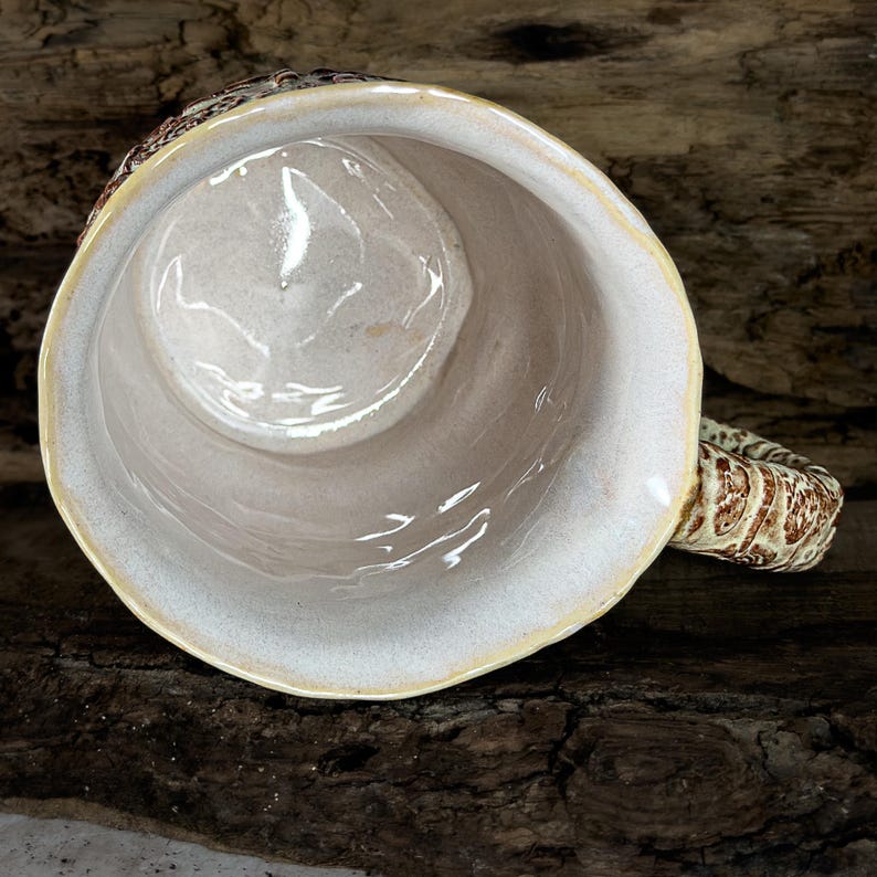 Op de afbeelding: Een keramische mok met een gladde, glanzende, gebroken witte binnenkant en een getextureerde, bruin en cr&egrave;mekleurige buitenkant. De mok heeft een dik, geribbeld handvat met een gevlekt patroon en een licht onregelmatige, organische rand.