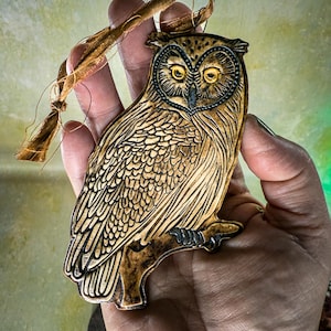 May include: A detailed, wooden owl ornament with a brown and black color scheme. The owl is perched on a branch and has yellow eyes. A brown string is attached for hanging. The ornament is held in a hand.