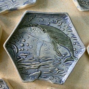 May include: Hexagonal ceramic plate with a raised design of a whale breaching the water. The plate is light blue with a darker blue whale and water details. Bubbles and water lines surround the whale.