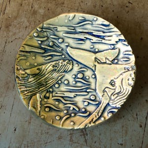 May include: A round ceramic plate with a whale design. The plate features a raised relief design of whales swimming in the ocean, with blue and yellow-green glaze. The design includes bubbles and water lines.