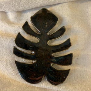 May include: A dark green ceramic leaf with a glossy finish and a light-colored edge. The leaf has a detailed, veined design and is set against a neutral background. This decorative piece is likely a home decor item.