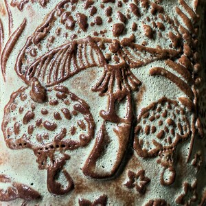 Op de afbeelding: Een keramisch oppervlak met een reli&euml;fontwerp van diverse paddenstoelen en kleine stervormen. De achtergrond is bleek, gedempt groen, terwijl de verhoogde paddenstoel- en stermotieven zijn afgewerkt met een contrasterend donkerbruin glazuur.