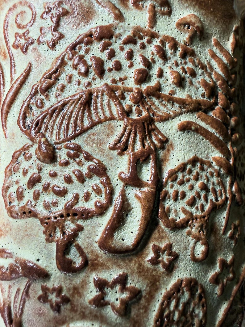 Op de afbeelding: Een keramisch oppervlak met een reli&euml;fontwerp van diverse paddenstoelen en kleine stervormen. De achtergrond is bleek, gedempt groen, terwijl de verhoogde paddenstoel- en stermotieven zijn afgewerkt met een contrasterend donkerbruin glazuur.