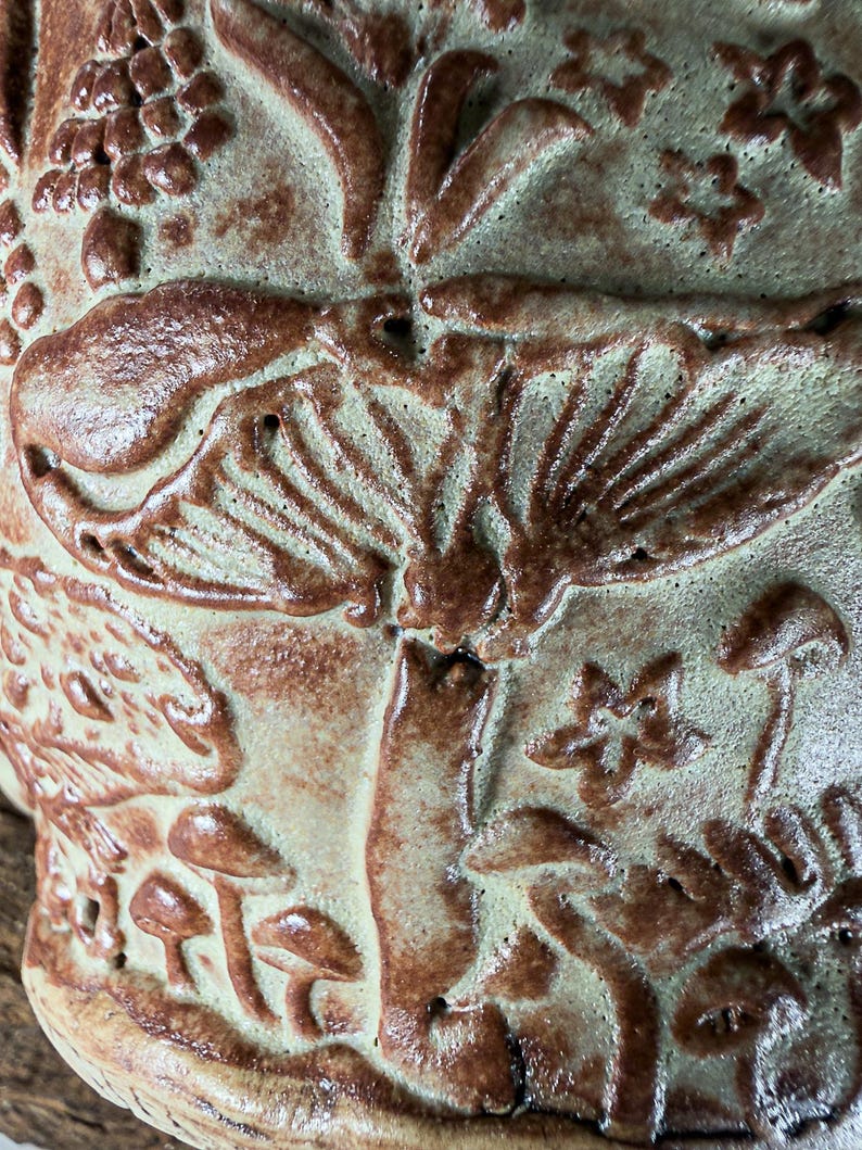 Op de afbeelding: Een keramieken vat met een reli&euml;fontwerp van diverse paddenstoelen en kleine stervormen. Het oppervlak heeft een licht saliegroene basiskleur met een donkerbruin glazuur dat de verhoogde paddenstoelhoeden, stelen en details accentueert.