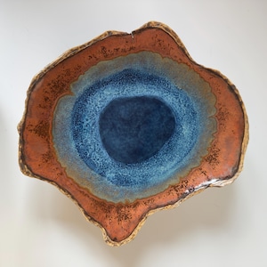 May include: A handcrafted ceramic bowl with a unique, organic shape. The bowl features a deep blue center, surrounded by layers of blue, gray, and rust-colored glaze. The rim has a textured, earthy appearance, adding to its artistic appeal.