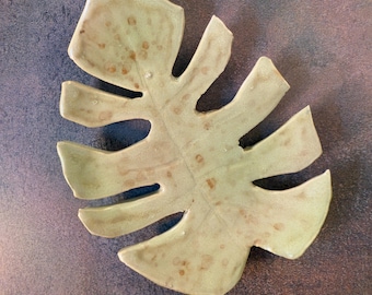 May include: A ceramic leaf-shaped dish with a light green glaze and brown speckles. The dish is shaped like a monstera leaf.