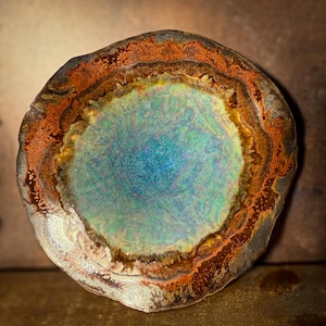 May include: A round, decorative ceramic piece with a textured surface. The center is a vibrant blue-green, surrounded by layers of yellow, orange, and brown. The outer edge is a dark, mottled gray. The piece has an organic, natural aesthetic.