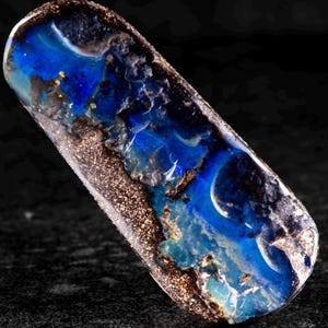 Könnte beinhalten: Ein polierter, rechteckiger Edelstein mit einem auffälligen Blau-Schwarz-Farbschema. Der Stein weist ein wirbelndes Muster aus tiefen Blautönen und Schwarz auf, mit Akzenten in Gold und Braun. Die Oberfläche hat eine strukturierte Erscheinung.