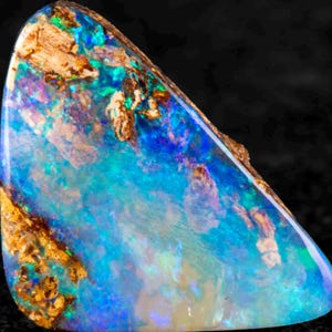 May include: A polished boulder opal gemstone featuring a vibrant play of colour with intense flashes of electric blue, green, and purple. The stone displays natural brown ironstone matrix inclusions embedded within the iridescent surface.