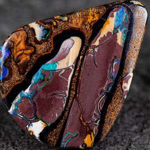 Puede incluir: Ópalo pulido de forma irregular que muestra una rica variedad de colores. La piedra presenta una base burdeos intenso, acentuada con negro, dorado y parches de azul iridiscente, verde y amarillo. El patrón único del ópalo se destaca por su superficie pulida.