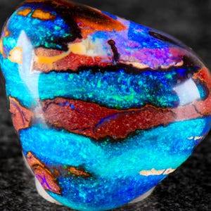Puede incluir: Una piedra preciosa de ópalo boulder pulida que presenta un juego de colores vibrante con bandas intensas de azul eléctrico, turquesa y rojo profundo. La superficie es lisa y redondeada, mostrando un patrón natural con inclusiones oscuras.