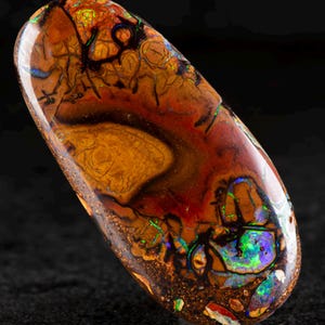 Puede incluir: Una piedra preciosa de ópalo pulida, de forma ovalada. La piedra muestra una rica paleta de colores, incluyendo tonos de marrón, rojo y toques de azul y verde. Se pueden ver patrones y texturas intrincados en la superficie.