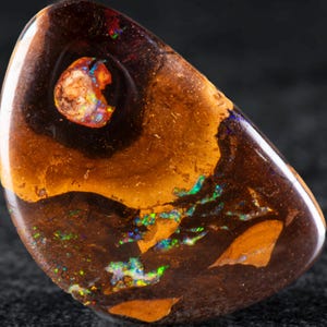 Könnte beinhalten: Ein polierter Boulder-Opal-Edelstein mit einer glatten, abgerundeten Dreiecksform. Der Stein zeigt eine dunkelbraune Eisensteinmatrix mit lebhaften, irisierenden Blitzen in Grün, Blau und Orange, die über die Oberfläche verteilt sind.