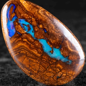 Puede incluir: Una piedra preciosa de ópalo pulida, de forma ovalada. El ópalo presenta un color base marrón terroso intenso con patrones y vetas intrincados. Una vibrante franja de azul y turquesa atraviesa el centro, creando un contraste llamativo. La piedra preciosa está sobre un fondo oscuro.