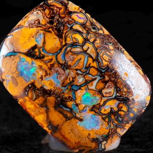 Puede incluir: Una gema rectangular pulida con un patrón vibrante de naranja, marrón, azul y blanco. La piedra tiene un diseño único y arremolinado con áreas de color iridiscente. La gema está sobre un fondo negro.