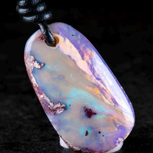 Puede incluir: Un colgante de ópalo de forma ovalada con una superficie lisa y pulida que presenta remolinos iridiscentes de color púrpura, azul y blanco, acentuados por inclusiones minerales oscuras. La piedra cuelga de un cordón negro grueso.