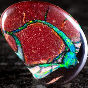 Könnte beinhalten: Ein polierter Boulder-Opal-Edelstein mit einer tief rotbraunen Eisensteinmatrix, durchzogen von lebendigen, irisierenden Adern aus elektrisch blauem und hellgrünem Opal, die auf der glatten, abgerundeten Oberfläche des Steins schimmern.