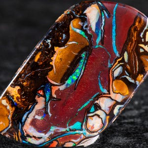 Peut inclure: Une pierre d'opale polie de forme ovale. La pierre présente une couleur de base rouge foncé avec des veines orange, marron, bleu et vert. L'opale a une finition très brillante, mettant en valeur ses motifs et couleurs internes.