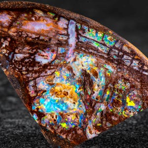 May include: A polished, triangular gemstone with a dark brown matrix and vibrant internal colours. The opal features flashes of blue, yellow, and green, creating a striking play of light. The stone's surface is smooth and reflective, showcasing its natural beauty.