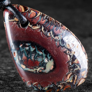 Peut inclure: Un pendentif en forme de goutte composé d'une pierre polie brun rougeâtre foncé avec des veines complexes et tourbillonnantes d'inclusions minérales irisées bleues, vertes et crème. Le pendentif est suspendu à un cordon en cuir noir.