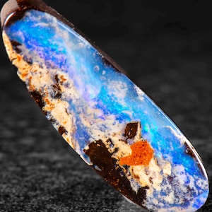 Könnte beinhalten: Ein länglicher, ovaler Boulder-Opal-Edelstein mit polierter Oberfläche, der lebhafte Blitze in elektrischem Blau und Weiß zeigt, kontrastiert durch eine dunkelbraune Eisensteinmatrix und einen kleinen, deutlichen Fleck aus orangefarbenen Einschlüssen.
