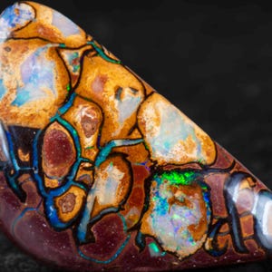 Może przedstawiać: Wypolerowany kamień szlachetny opal boulder o trójkątnym kształcie, z ciemnobrązową matrycą żelazistą przeplataną żywymi, opalizującymi błyskami niebieskiego, zielonego i złotego koloru na całej teksturowanej, organicznej powierzchni.