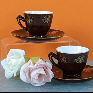 Könnte beinhalten: Zwei braune Teetassen mit goldenen Blumendekoren und weißem Innenraum, jede auf einer passenden Untertasse mit goldenen Blumendekoren. Die Untertassen sind auf einem klaren Acryl-Präsentationsständer gestapelt.