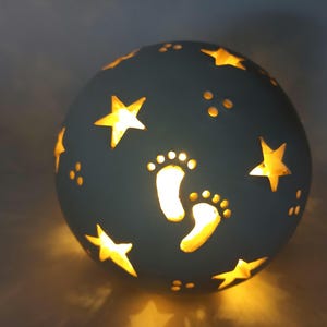 Könnte beinhalten: Eine kugelförmige Nachtlampe in Türkis mit ausgeschnittenen Sternen- und Fußabdruck-Designs. Das Innere leuchtet mit warmem, gelbem Licht und erzeugt eine sanfte Atmosphäre. Die Lampe ist ein dekorativer Artikel, ideal für ein Kinderzimmer.