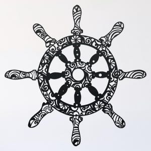 May include: Black and white decorative ship's wheel with intricate floral designs. The wheel has eight spokes and a circular center.
