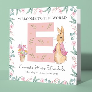 May include: A pink and white card with a floral border. The card says "Welcome to the world" and features a pink letter "E" with a small rabbit illustration. The card is personalised with the name "Emmie Rose Teasdale" and the date "Thursday 13th December 2023".