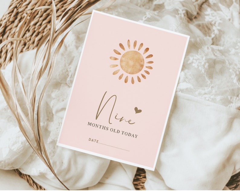 Pu&ograve; includere: Un biglietto rosa con un'illustrazione di sole dorata e il testo "Nine months old today" con una riga vuota per la data.
