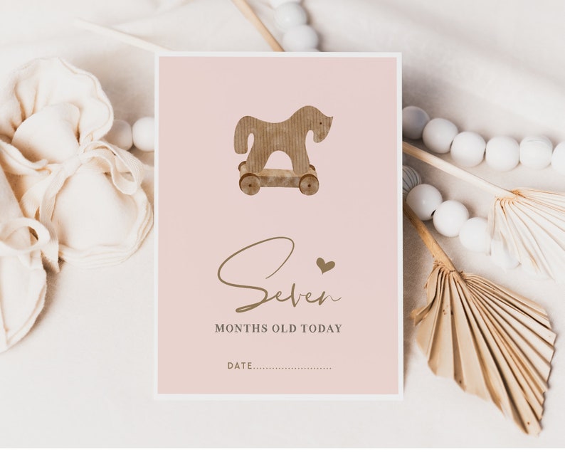 Pu&ograve; includere: Un biglietto rosa con un'illustrazione di un cavallo a dondolo di legno marrone e il testo "Seven months old today" in lettere dorate. Il biglietto ha uno spazio vuoto per una data.