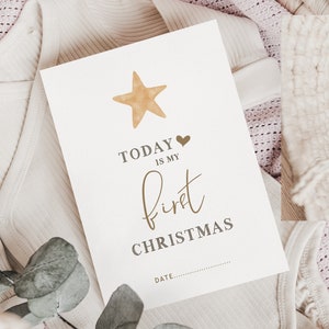Pu&ograve; includere: Un biglietto bianco con il testo "TODAY IS MY first CHRISTMAS" e una stella dorata con un cuore sopra il testo. Il biglietto &egrave; su un tessuto a maglia bianco e rosa.