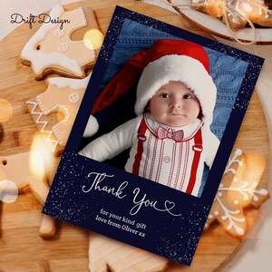 May include: A navy blue Christmas card with a white snowflake pattern. The card features a photo of a baby wearing a red Santa hat and a red and white striped outfit. The text "Thank You" is written in white cursive with a heart symbol. The text "for your kind gift love from Oliver xx" is written in white below the photo.