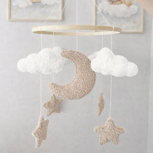 Puede incluir: Móvil para bebé con un aro de madera, una luna creciente beige, nubes blancas y estrellas beige. El móvil está suspendido, con dos imágenes enmarcadas de un oso y un conejo sobre nubes en el fondo.