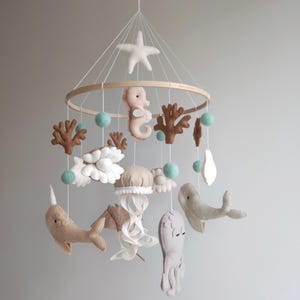 Op de afbeelding: Een babymobiel van vilt met een nautisch thema. Het heeft een ster, een zeepaardje, een kwal, een octopus, een walvis, een narwal en koraal. De mobiel hangt aan een houten ring en heeft lichtblauwe pompoenen.