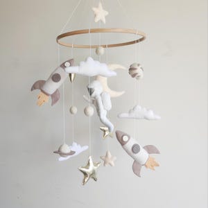 Op de afbeelding: Een witte, grijze en gouden vilt babymobiel met een raket, een astronaut, wolken, sterren en planeten. De mobiel hangt aan een houten ring.