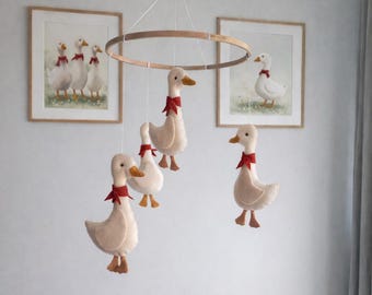 Op de afbeelding: Een baby mobile met vijf crème-kleurige pluche ganzen, elk met een rode strik, hangend aan een houten ring. Twee ingelijste prenten van ganzen met rode strikken hangen aan de muur. De ganzen hebben oranje snavels en poten.