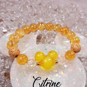 May include: Citrine gemstone bracelet with a yellow Mickey Mouse charm. The bracelet is made with natural citrine stones and lava beads. The bracelet is on a white background with glitter.