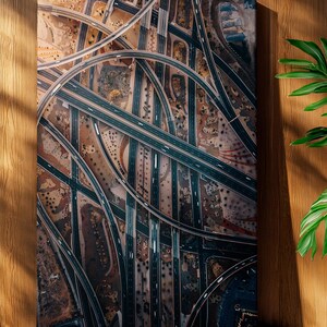 May include: An aerial view of a complex highway interchange, printed on canvas. The artwork features numerous curving roads and overpasses in shades of brown, gray, and black. The canvas is displayed against a wooden wall with a green plant in the corner.