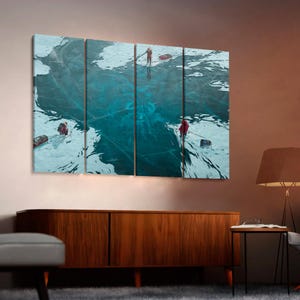 May include: A four-panel wall art set depicting a vast, cracked frozen lake surface in deep teal and white. Three figures in red winter gear are scattered across the ice, each pulling a small sled, with a large dark shape visible beneath.