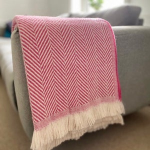 Op de afbeelding: Een roze en witte herringbone deken met een franje rand. De deken is over een grijze bank gelegd.